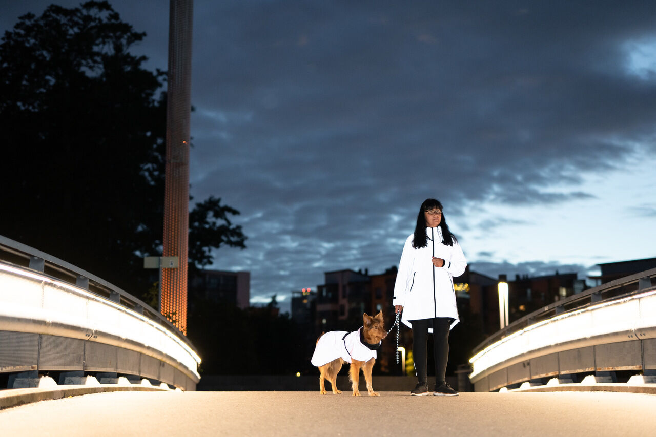 PAIKKA-Regenmantel-fuer-Zweibeiner-reflektierend-Human-Visibility-Raincoat-Frau-spaziert-auf-Bruecke-mit-Hund-60-46253